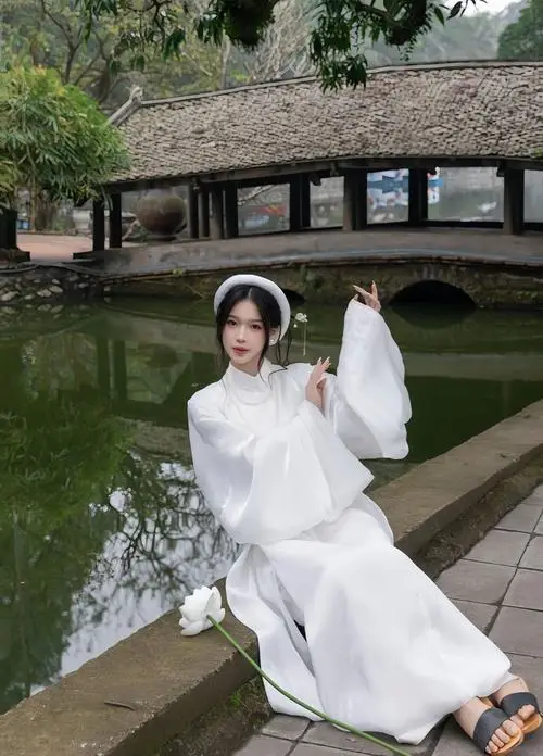 Cô gái áo dài trắng ngồi bên hồ nước cổ, tay nâng nhẹ vành nón lá, tà áo phủ mềm trên bậc đá