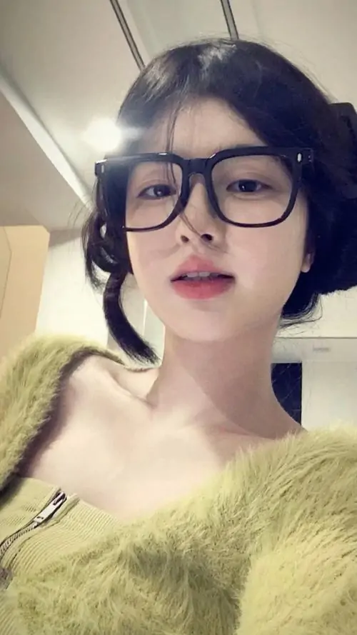 Cô gái tóc đen đeo kính nhìn thẳng vào máy, áo lông vàng nhạt tạo cảm giác mềm mại trong khung ảnh selfie sáng nhẹ