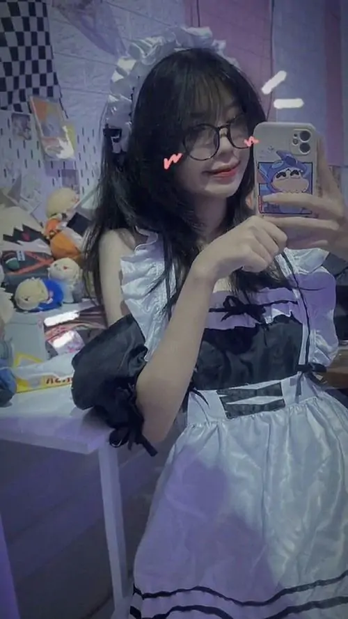 Dưới ánh đèn xanh tím, cô gái đeo kính ngồi trong phòng, váy trắng và mái tóc dài tạo cảm giác ảnh mỹ nhân mạng xã hội dịu mắt