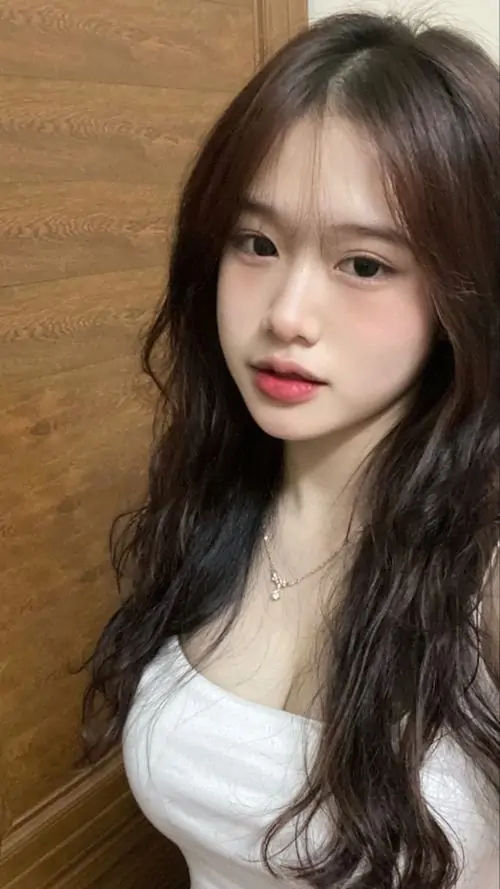 Ánh sáng trong phòng làm rõ gương mặt thanh tú của cô gái tóc dài, áo trắng đơn giản gợi cảm giác ảnh gái đẹp mạng xã hội hiện đại