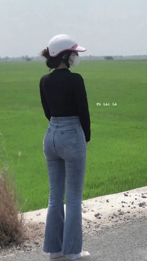 Cánh đồng xanh phía xa làm nền cho cô gái áo đen, quần jean ống loe ôm, đội mũ trắng và đứng quay lưng thư thái