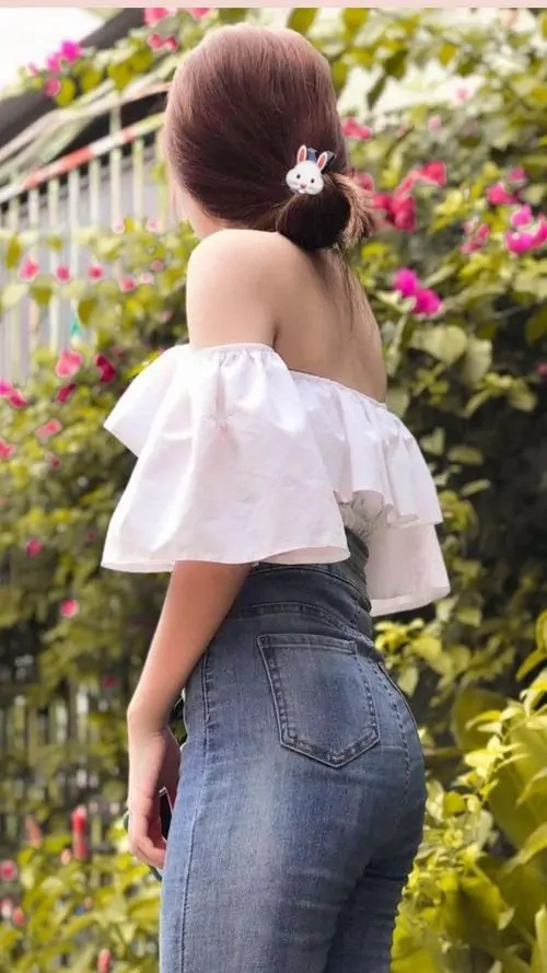 Giàn hoa rực rỡ phía sau làm nổi bật cô gái áo trễ vai trắng, quần jean xám ôm, tóc búi thấp nhìn nghiêng dịu dàng