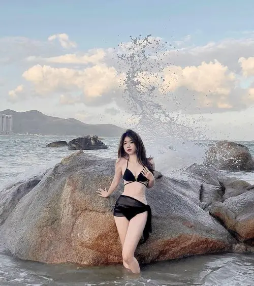 Sóng biển tung bọt sau lưng cô gái mặc bikini đen, dáng ngồi trên đá và nụ cười nhẹ làm bức ảnh mùa hè thêm sống động