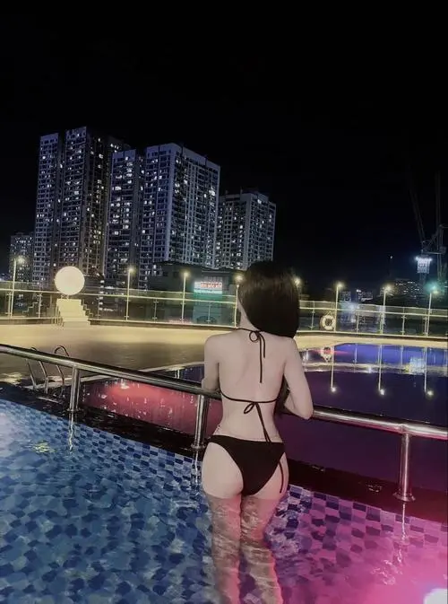 Ảnh hot girl mặc bikini đen quyến rũ chụp từ sau bên hồ bơi đêm, ánh đèn thành phố phản chiếu mặt nước tạo tông màu nổi bật