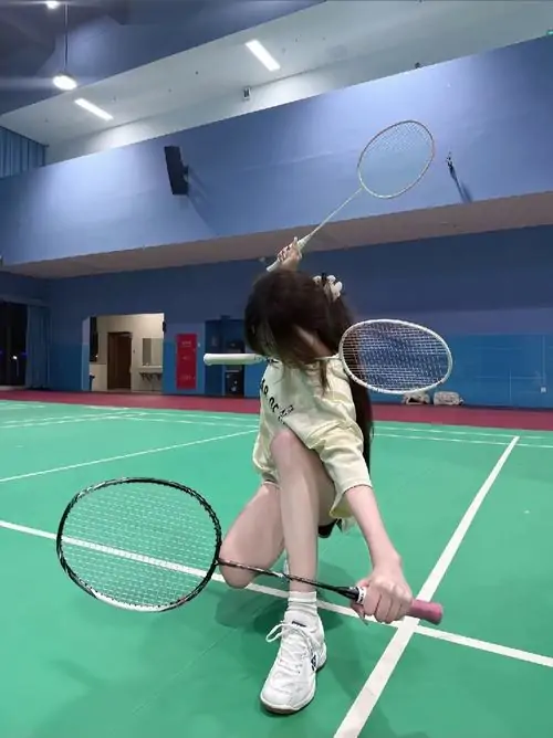 Cô gái ngồi trên sân cầu lông trong nhà, tay cầm vợt sau lưng, áo trắng và váy ngắn tạo phong cách thể thao nữ tính