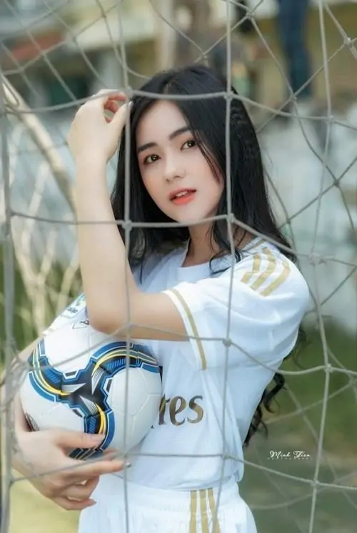 Qua lớp lưới sân bóng, cô gái ôm quả bóng trước ngực, áo đấu trắng xanh và mái tóc đen dài tạo ánh nhìn trong trẻo