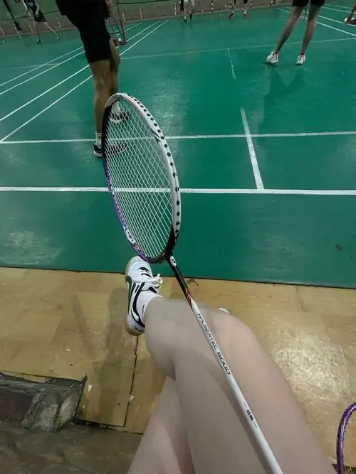 Góc cận cảnh vợt tennis đặt trên nền sân xanh, giày thể thao trắng và chân người chơi xuất hiện nhẹ phía sau