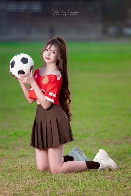Cô gái quỳ trên sân cỏ, ảnh người đẹp yêu bóng đá mặc áo đỏ phối váy nâu, tay cầm bóng và cười tươi trước ống kính