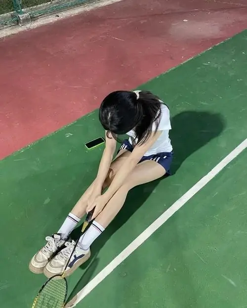 Cô gái ngồi cúi người buộc dây giày trên sân thể thao, áo trắng xanh và váy ngắn tạo cảm giác chuẩn bị vận động