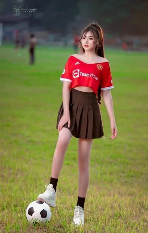 Áo đấu đỏ nổi bật trên nền sân cỏ tối, cô gái đứng cạnh quả bóng, tóc nâu buông vai và gương mặt rạng rỡ