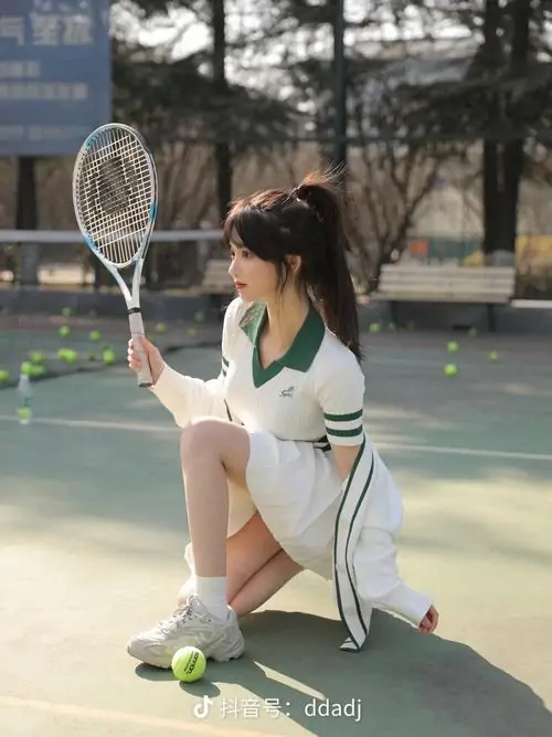 Cô gái ngồi trên sân tennis cạnh nhiều bóng vàng, áo trắng phối chân váy cùng tóc buộc cao tạo dáng năng động
