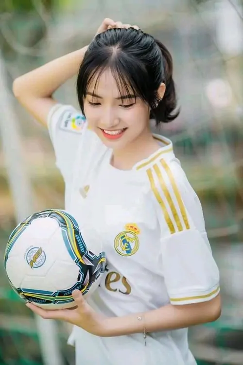 Cô gái ôm bóng trước ngực trong áo Real Madrid trắng, tóc nâu búi thấp, nụ cười tươi làm khung ảnh rất gần gũi