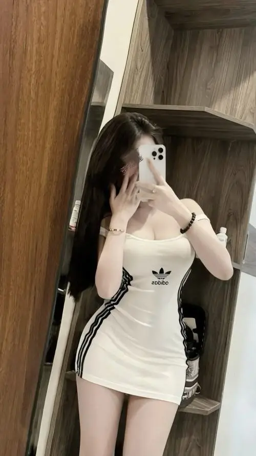 Cô gái có mái tóc đen dài diện bộ váy ngắn màu trắng ôm sát cơ thể khoe trọn đôi chân thon dài bên bức tường gỗ trầm ấm