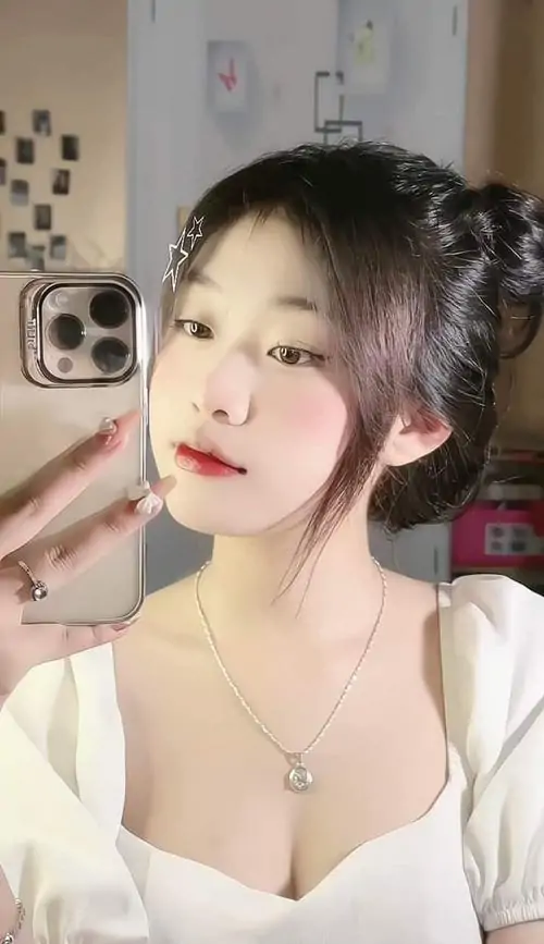 Góc selfie cận mặt cho thấy cô gái gái xinh đẹp nhất tóc búi cao, áo trắng cổ mềm và ánh nhìn nhẹ dưới nền phòng sáng