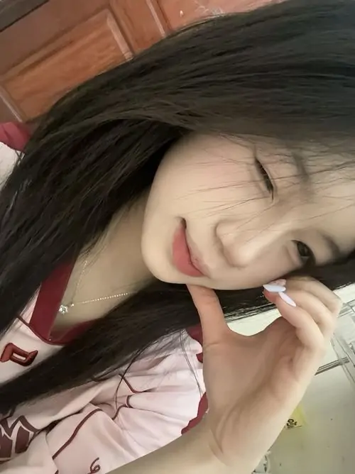 Một cô gái tóc ngắn cười nhẹ khi cầm điện thoại, áo len trắng và nền phòng tối tạo cảm giác ảnh thiếu nữ mạng xã hội gần gũi