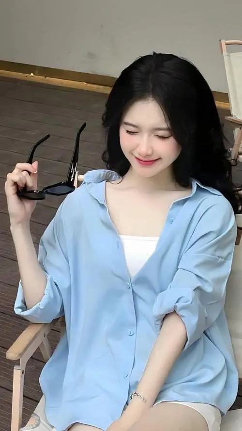 Áo xanh nhạt cổ rộng giúp cô gái ngồi trước gương trông mềm mại, mái tóc đen buông dài và nụ cười kín đáo rất nhẹ