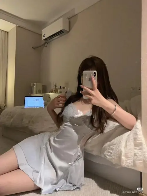 Trước gương phòng ngủ, cô gái mặc áo choàng ngủ trắng cầm điện thoại, ánh đèn vàng làm tổng thể thêm dịu mắt