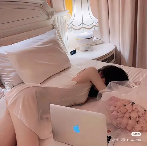 Giường hồng và laptop mở phía trước tạo bối cảnh đời thường, cô gái nằm sấp trong đồ ngủ sáng màu rất thoải mái
