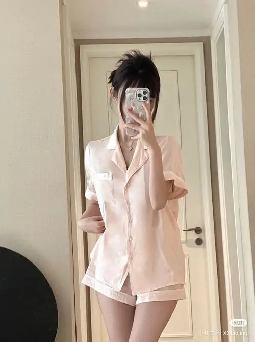 Cô gái mặc áo choàng hồng đứng trước gương, tay cầm điện thoại che mặt, mái tóc dài phủ ngang vai mềm mại