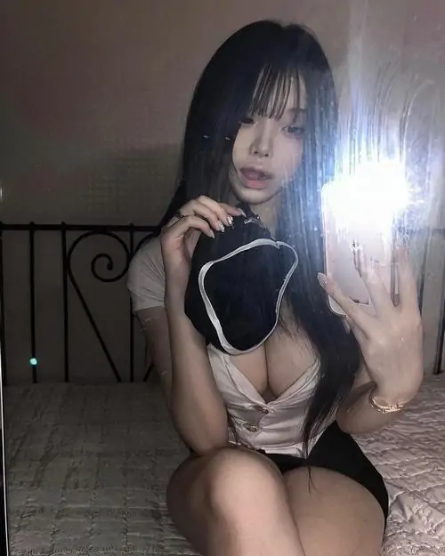 Cô gái tóc ngắn ngồi trước gương với váy ngủ đen trắng, tay cầm điện thoại và biểu cảm che mặt đầy tinh nghịch