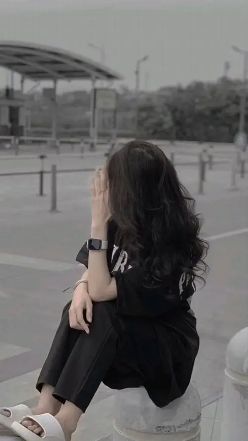 Không gian ngoài trời hơi mờ, gái đẹp che mặt trong áo đen cầm túi nhỏ, mái tóc xoăn dài nổi bật trên nền xám nhạt