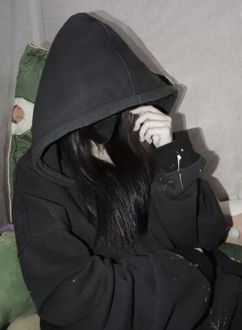 Hoodie đen trùm kín đầu, cô gái cúi mặt giữa nền phòng sáng, tay áo che gần hết khuôn mặt tạo bố cục kín đáo