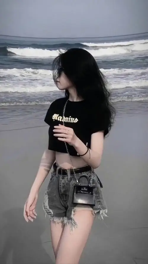 Trên bãi biển nhiều gió, cô gái mặc áo đen và quần short rách đi nghiêng, tóc dài che mặt tạo cảm giác rất đời thường