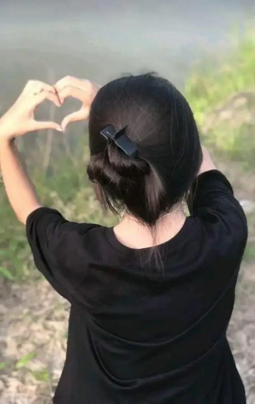 Từ phía sau, cô gái áo đen buộc tóc thấp giơ hai tay chỉnh tóc, nền cây xanh làm bức ảnh có màu tự nhiên