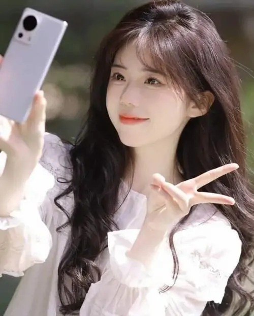 Cô gái áo trắng ren cầm điện thoại selfie, tóc nâu uốn lọn và dáng tay chữ V tạo cảm giác nhẹ nhàng đời thường