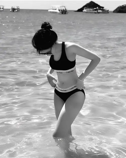 Cô gái bước nhẹ trên cầu gỗ ven biển với áo choàng trắng ngoài bikini đen, gió thổi mái tóc tạo cảm giác thư thái
