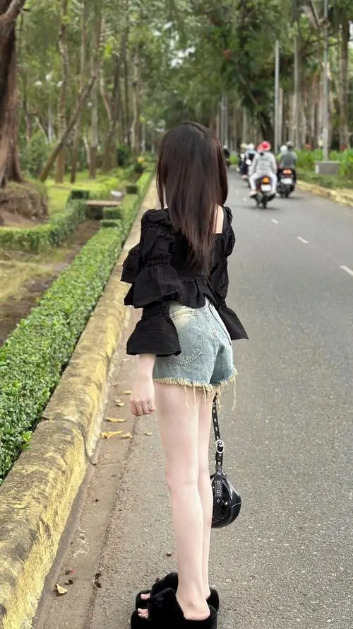Tường gạch nâu làm nền cho cô gái áo thun be, quần jean đen bó, tóc xoăn dài và dáng đứng tự tin