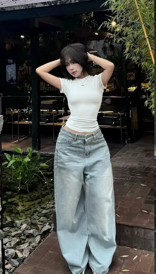 Trong studio nền trắng, hình người đẹp phối quần jean với áo croptop đen, thắt lưng nhỏ và dáng ngồi nghiêng thanh thoát