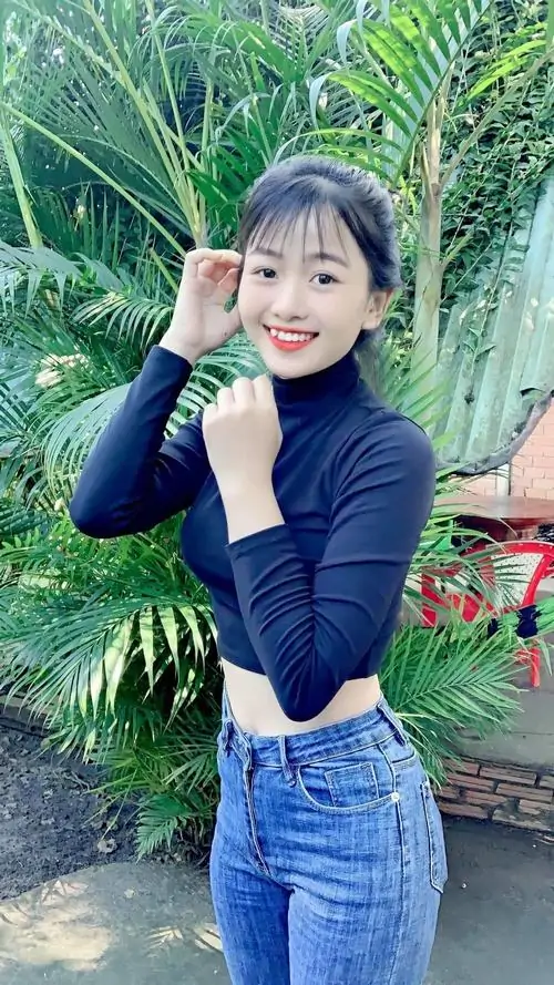 Nền lá nhiệt đới xanh mướt làm cô gái áo len xanh, jean xanh cạp cao và mái tóc ngắn mỉm cười trông tươi tắn