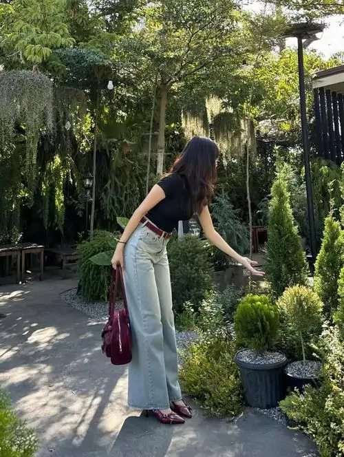 Khu vườn nhiều chậu cây tạo nét yên bình cho cô gái áo đen, quần jean xanh, túi đỏ và dáng cúi nhẹ chăm cây