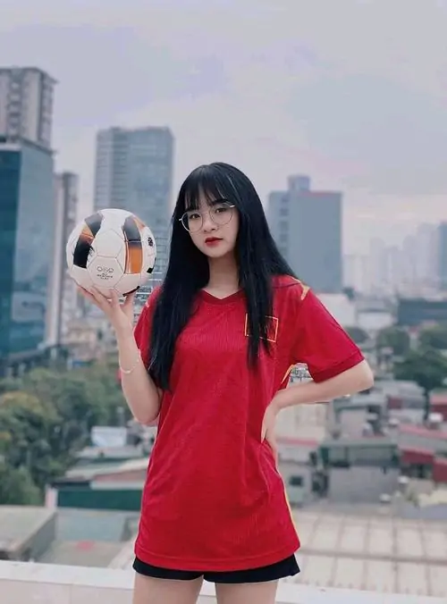 Cô gái mặc áo Real Madrid trắng ngồi bên lưới khung thành, tay giữ quả bóng, nụ cười nhỏ tạo cảm giác thân thiện