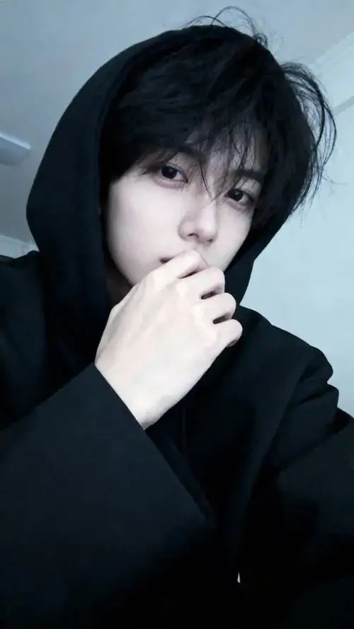 Không gian phòng tối làm nổi bật một ảnh nam tử hán mặc áo hoodie đen đang đưa tay lên chạm nhẹ vào cằm với biểu cảm suy tư đầy cuốn hút