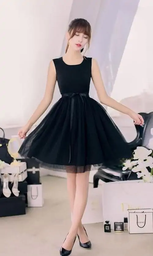 Đường may tinh tế ôm eo giúp chiếc đầm đen gọn dáng, cô gái cầm túi nhỏ giữa khung nền studio sáng