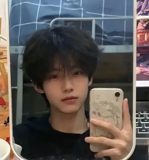 Chàng trai trẻ che khuyết một phần gương mặt bằng chiếc điện thoại khi chụp ảnh selfie trong phòng ngủ khoe mái tóc đen dày dặn được cắt tỉa gọn gàng
